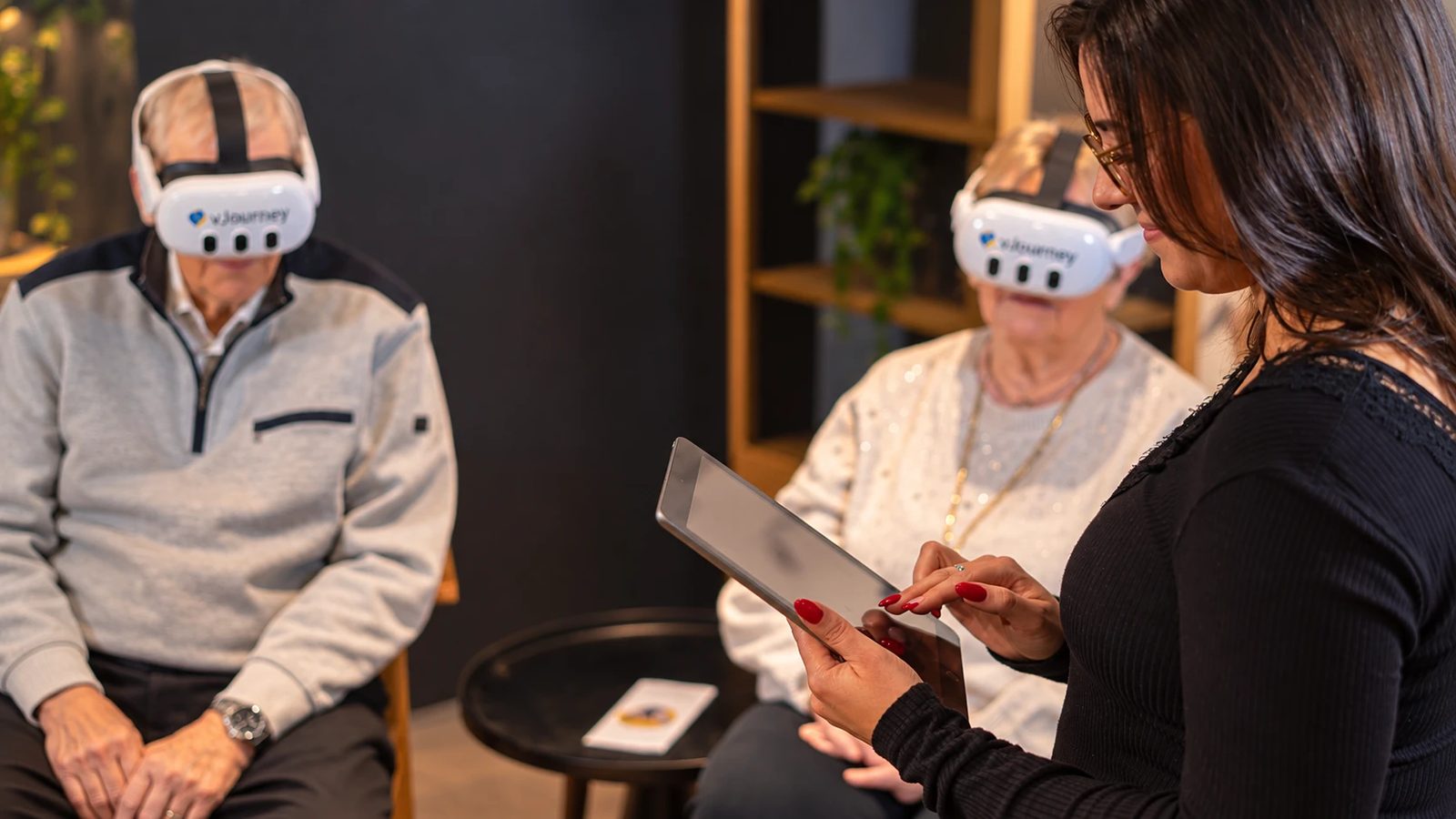 Zwei Senioren mit vJourney-VR-Brillen, eine Betreuerin steuert die Session per Tablet